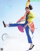 A woman in a colorful jacket and blue pants is jumping in the air.